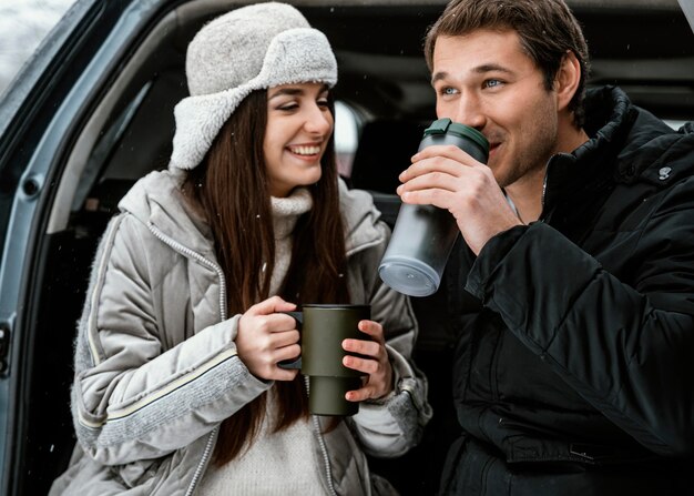 Front view of smiley couple having a warm drink in the car's trunk while on a road trip