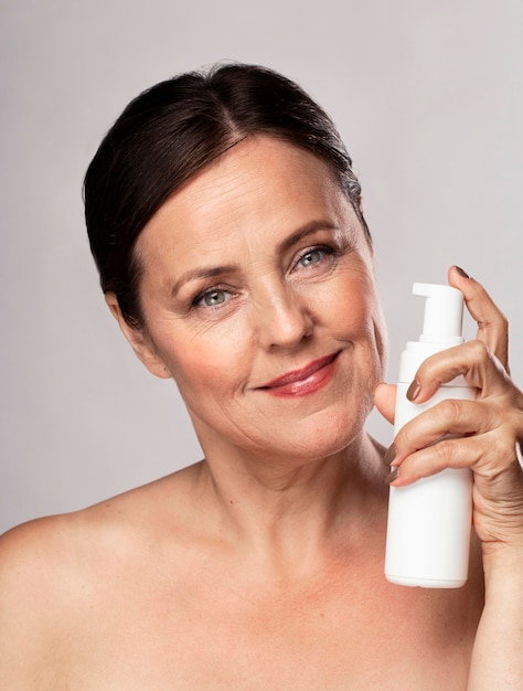 Front view of smiley elder woman holding bottle of cleanser for skincare