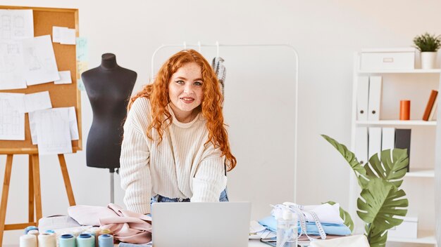 Front view of smiley female fashion designer working in atelier with laptop