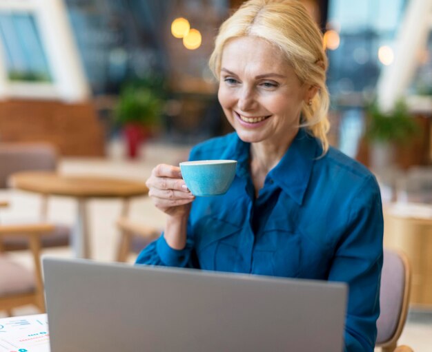 Front view of smiley older business woman having cup of coffee and working on laptop