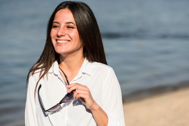 Front view of smiley woman on the beach holding sunglasses