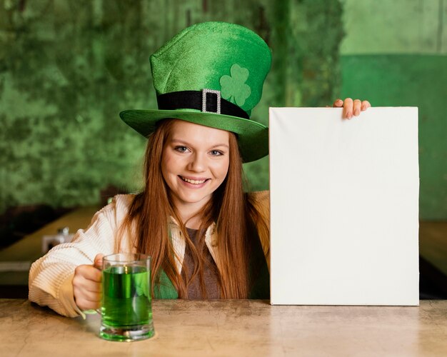 Front view of smiley woman celebrating st. patrick's day at the bar with blank placard and drink