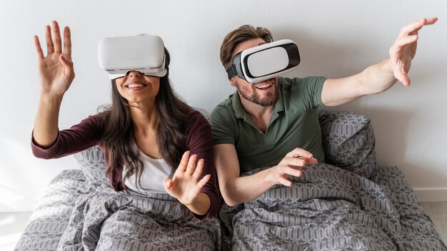 Front view of smiley woman and man using virtual reality headset in bed