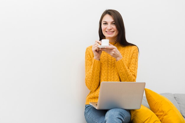 ソファの上に座って、ラップトップとラップのコーヒーのカップを保持している笑顔の女性の正面図