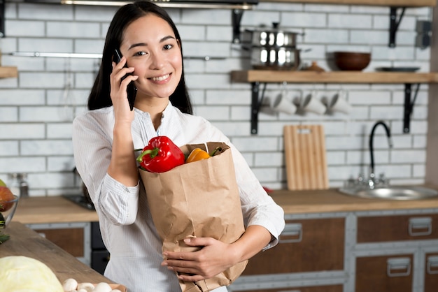 Front view of smiling asian woman talking on mobile phone while holding grocery bag