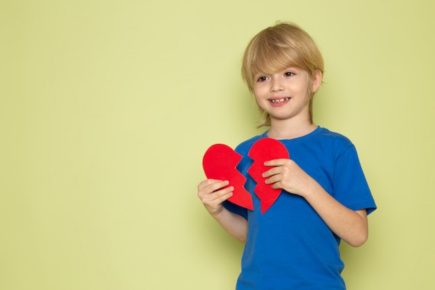 A front view smiling blonde boy in blue t-shirt holding teared heart shape on the stone colored backgorund