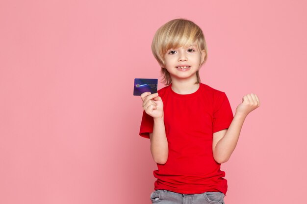 ピンクの床に赤いtシャツに紫のカードを保持している金髪の少年の笑顔正面図