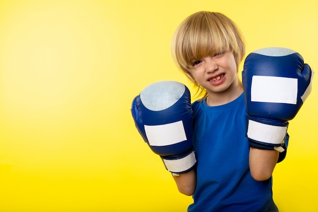 Вид спереди улыбающегося белокурого мальчика, позирующего бокс в синих боксерских перчатках на желтой стене