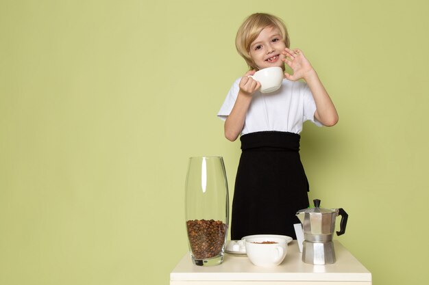 sotne色の机の上のテーブルの近くのコーヒーを準備する白いtシャツで金髪の少年の笑顔正面図