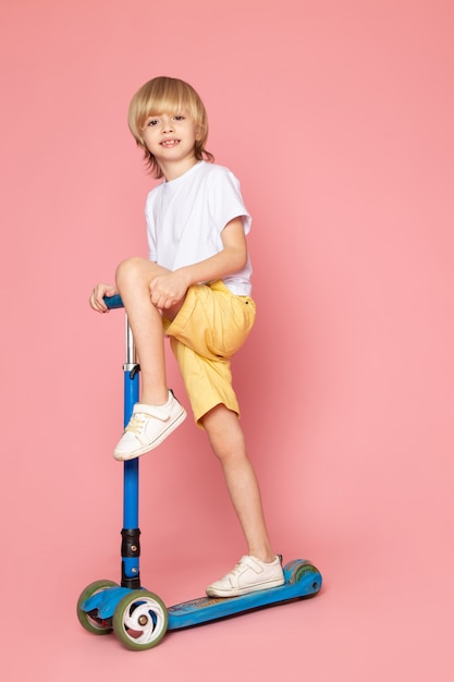 A front view smiling blonde boy in white t-shirt riding scooter on the pink space