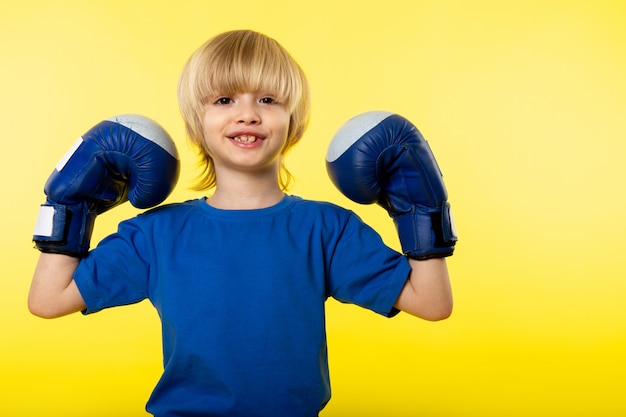 A front view smiling blonde flexing with blue boxing gloves on the yellow wall