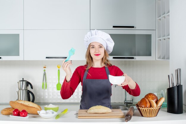 Front view smiling blonde woman in cook hat and apron holding brush and bowl in the kitchen