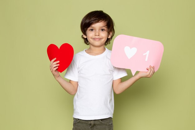 A front view smiling boy adorable sweet in white t-shirt holding heart shape and white sign on the stone colored space