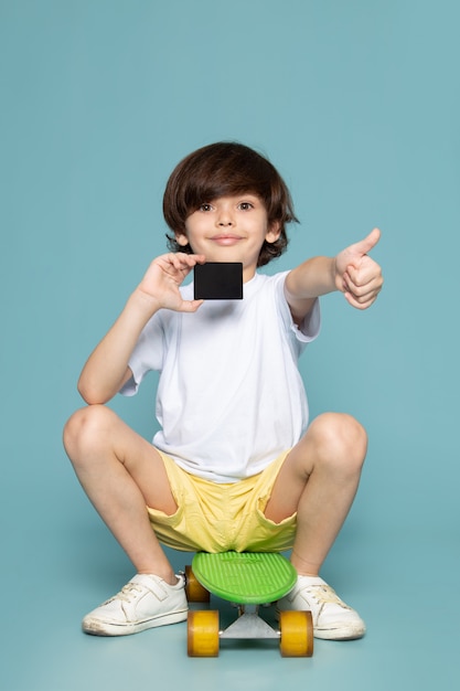 A front view smiling boy in white t-shirt and yellow jeans riding green skateboard on the blue space