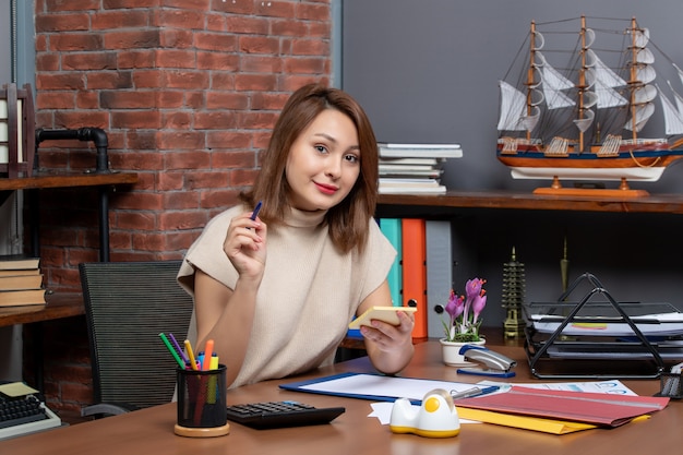 Front view of smiling business woman holding pen and note papers sitting at wall