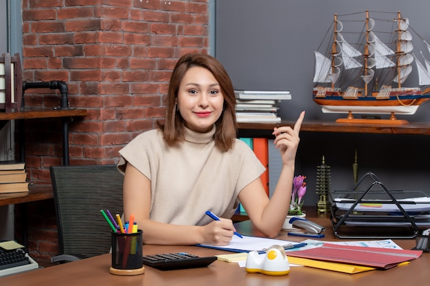 Front view of smiling business woman pointing finger up sitting at wall