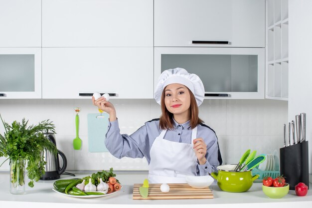 Front view of smiling chef and fresh vegetables with cooking equipment and holding eggs in the white kitchen