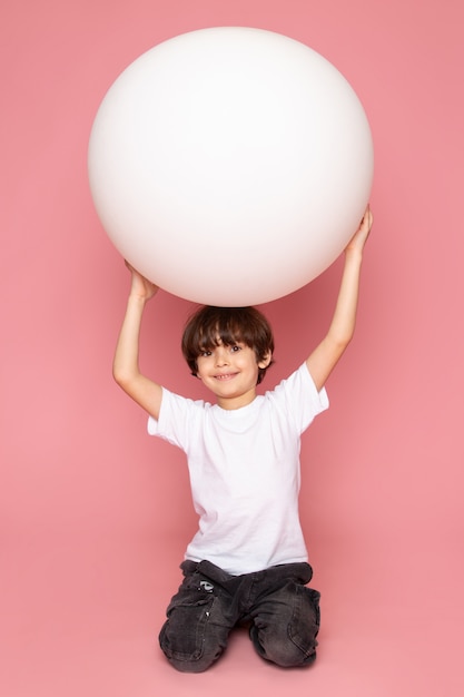 A front view smiling child boy in white t-shirt playing with white ball on the pink space