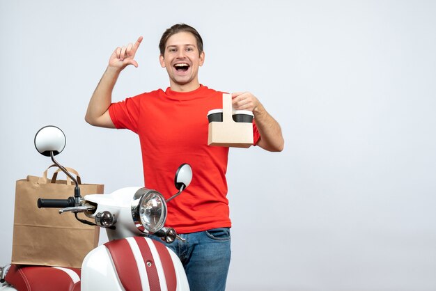 Front view of smiling confident happy delivery man in red uniform standing near scooter showing order on white background