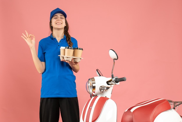 Вид спереди улыбающейся курьерской девушки, стоящей рядом с мотоциклом, держащей в руках кофе, делая жест очки на фоне пастельного персикового цвета