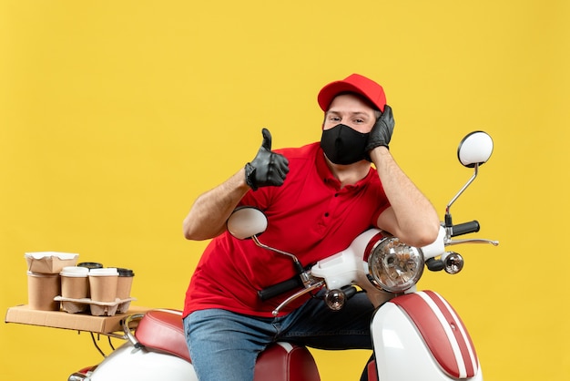 Front view of smiling courier man wearing red blouse and hat gloves in medical mask delivering order sitting on scooter making ok gesture