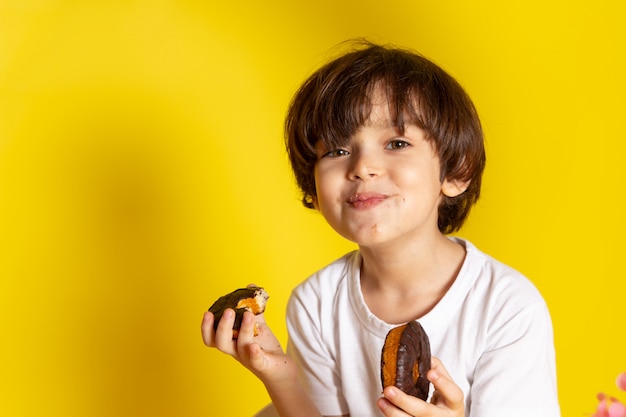 黄色の机の上の白いtシャツでチョコドーナツを食べて笑顔のかわいい男の子の正面図