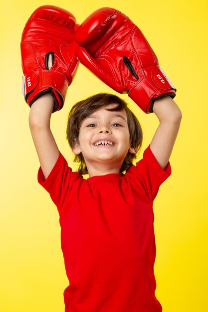 A front view smiling cute kid in red t-shirt and red boxing gloves on the yellow wall