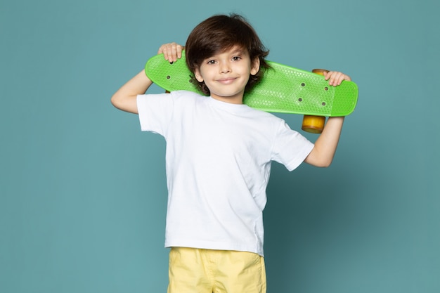 A front view smiling cute kid in white t-shirt holding skateboard on the blue floor