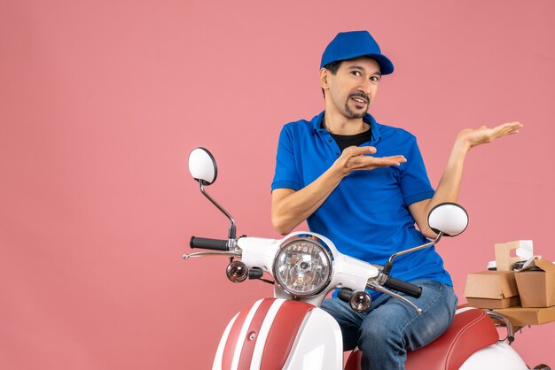 Front view of smiling delivery guy wearing hat sitting on scooter and pointing something on the left side on pastel peach background