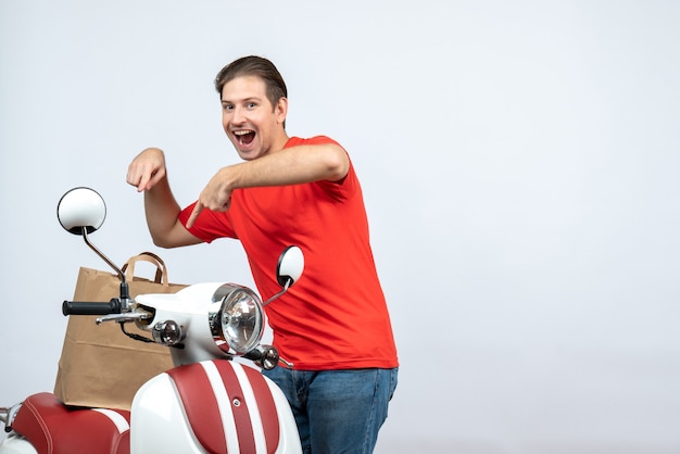 Front view of smiling delivery man in red uniform standing near scooter pointing order on white background