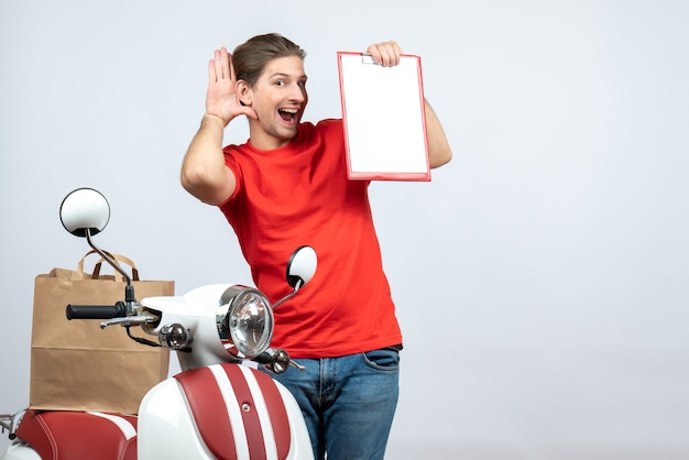 Front view of smiling delivery man in red uniform standing near scooter showing document listening to the last gossiping on white background