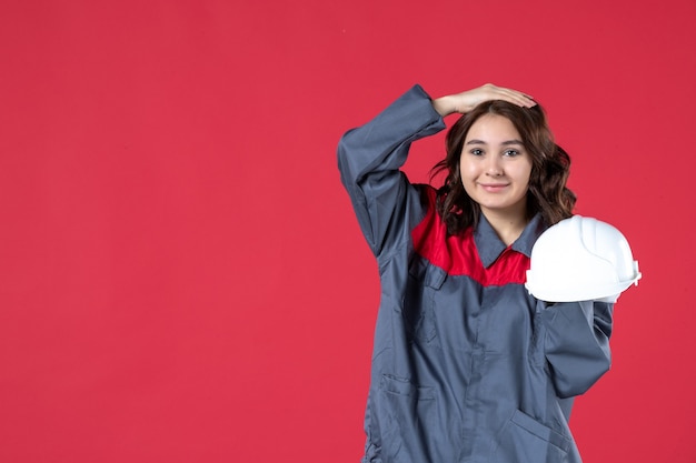 Vista frontale dell'architetto femminile sorridente che tiene il cappello duro e che mette la mano sulla sua testa su fondo rosso isolato