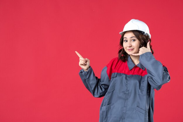 Front view of smiling female builder in uniform with hard hat and making call me gesture pointing up on isolated red background