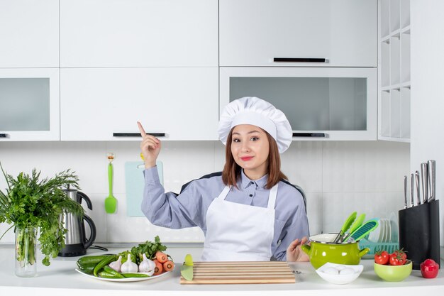 Front view of smiling female chef and fresh vegetables pointing up on the right side in the white kitchen