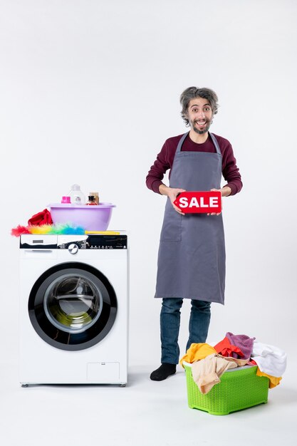 Front view smiling guy in apron holding up sale sign standing near washer on white wall