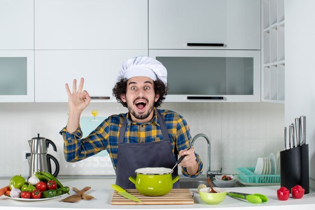 Front view of smiling and happy male chef with fresh vegetables tasting ready meal and making eyeglasses gesture in the white kitchen