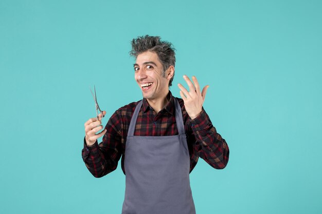 Front view of smiling happy male hairdesser wearing gray apron and holding scissor on blue soft color background