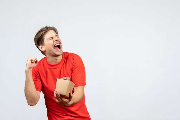 Front view of smiling happy young guy in red blouse holding small box on white background