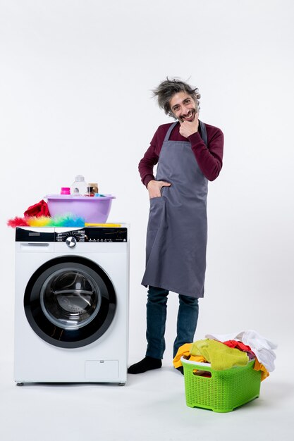 Front view smiling housekeeper man in apron standing near white washing machine on white wall