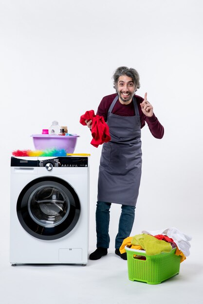 Front view smiling housekeeper man holding red towel standing near washing machine on white wall