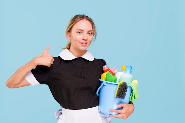 Front view of smiling janitor woman showing thumb up sign while holding cleaning products in bucket