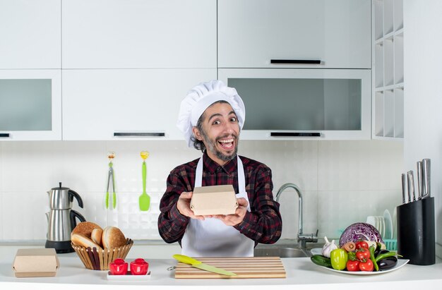 Front view of smiling male chef holding box standing behind kitchen table in the kitchen