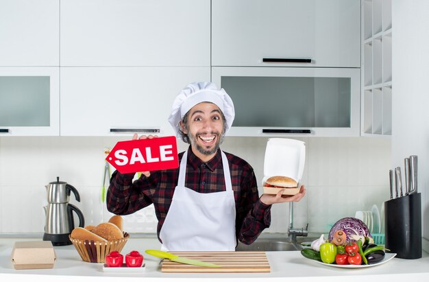 Front view of smiling male chef holding up sale sign and burger in the kitchen