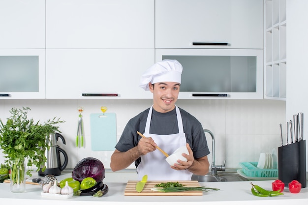 Front view smiling male chef in uniform holding bowl and spoon behind kitchen table