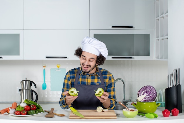 Вид спереди улыбающегося шеф-повара со свежими овощами, готовящего с кухонными принадлежностями и держащего нарезанный зеленый перец на белой кухне