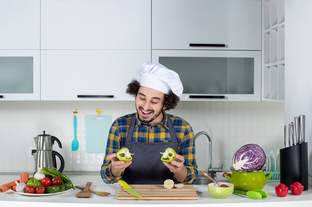 Front view of smiling male chef with fresh vegetables and cooking with kitchen tools and holding the cut green peppers in the white kitchen