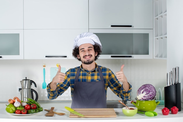 Front view of smiling male chef with fresh vegetables and cooking with kitchen tools and making ok gesture in the white kitchen