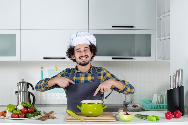 Вид спереди улыбающегося шеф-повара со свежими овощами, указывающего на себя и еду на белой кухне