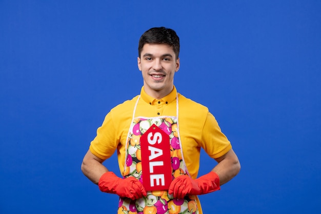 Free photo front view of smiling male housekeeper in yellow t-shirt holding sale sign on blue wall