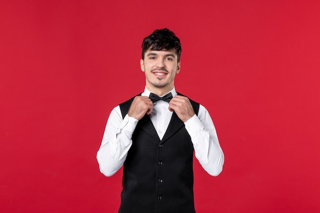 Front view of of smiling male waiter in a uniform with bowtie on neck on red wall